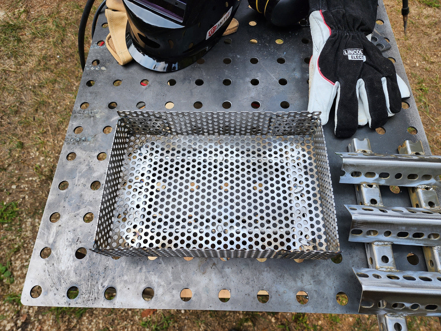 One large perforated steel basket and one square tube and angle iron wood pellet insert on a welding table