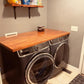 Butcher block countertop for laundry room folding and utility upgrade over front-loading washer and dryer, with a wall shelf holding cleaning supplies.