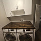Custom butcher block countertop for laundry room over front-load washer and dryer, dark wood with white cabinets.