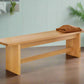 A white oak wood bench with a cushion on it, placed against a light blue wall on a hardwood floor.